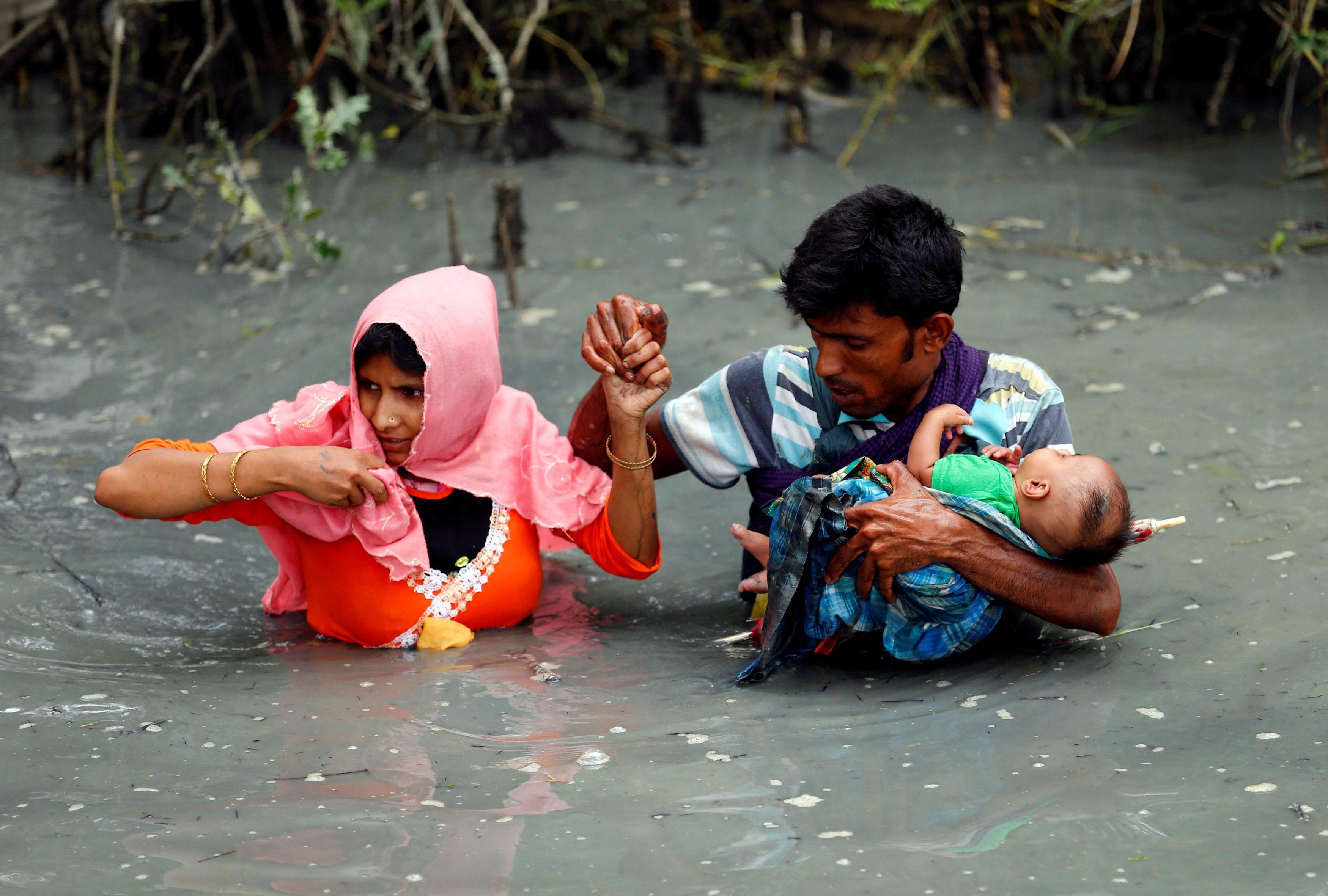Rohingya refugees carry their child as they walk through water after crossing border by boat through