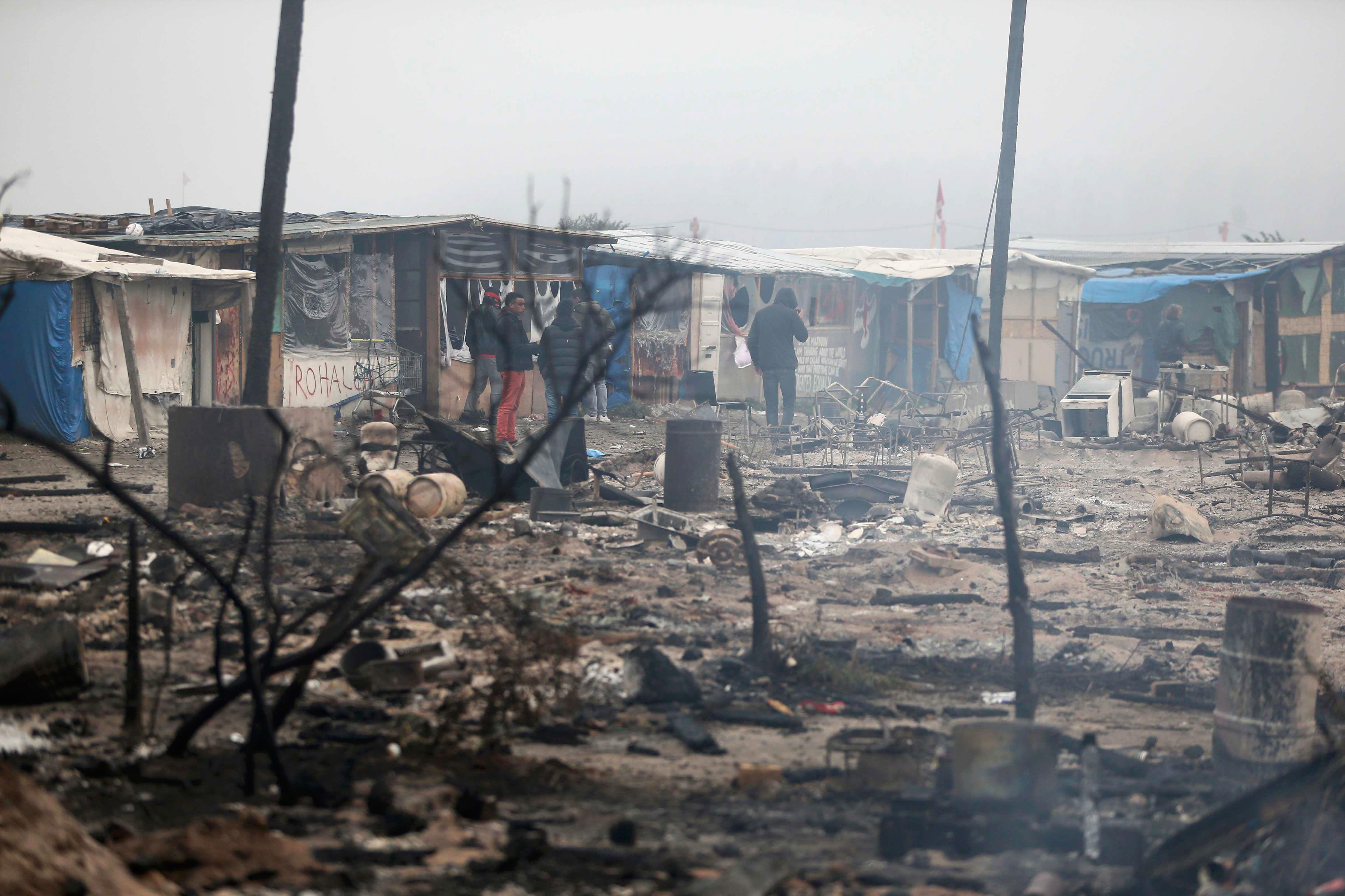 Miigrants stand in the charred debris from destroyed makeshift shelters and tents in the 