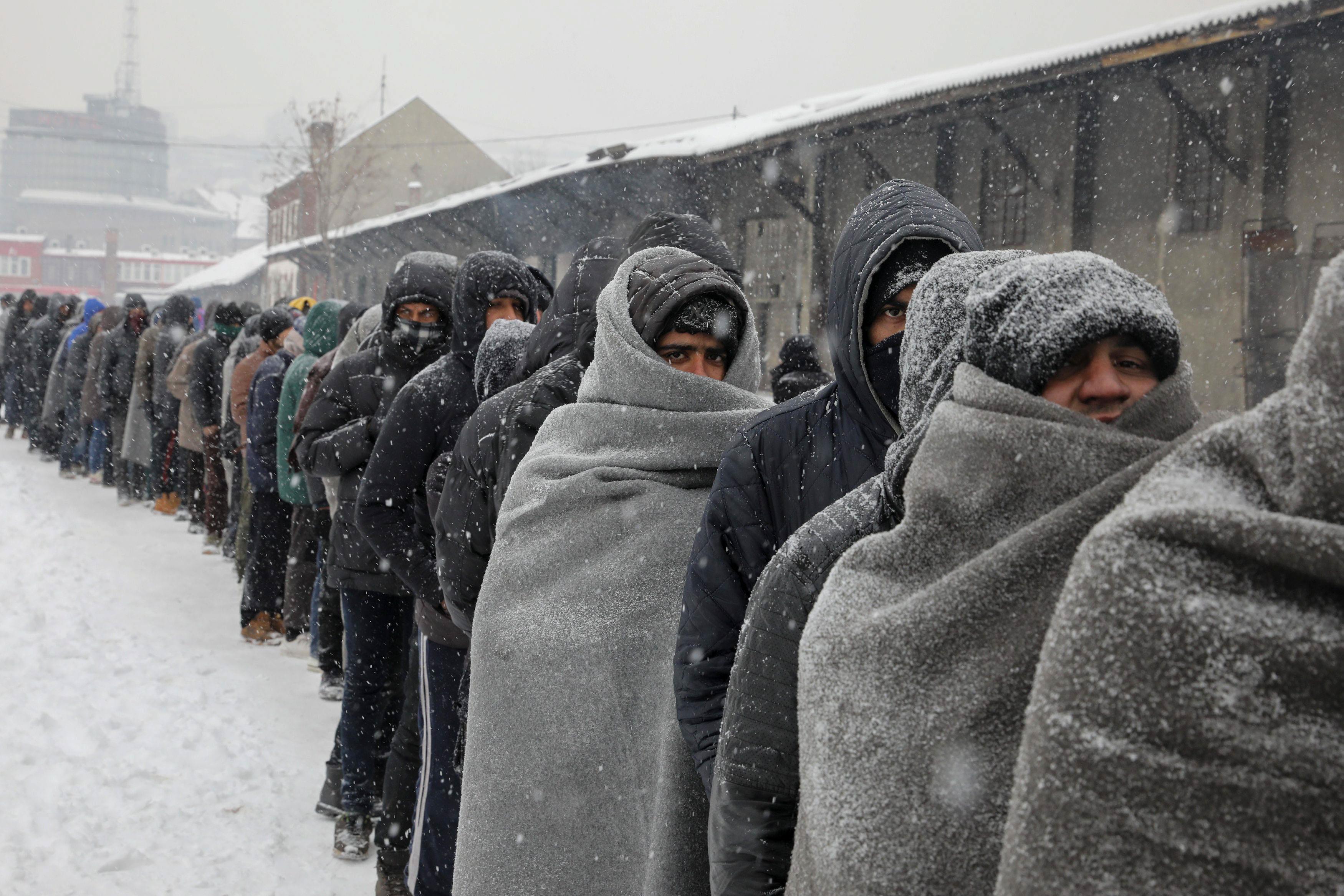 Migrants wait in line to receive a plate of free food during a snowfall outside a derelict customs w