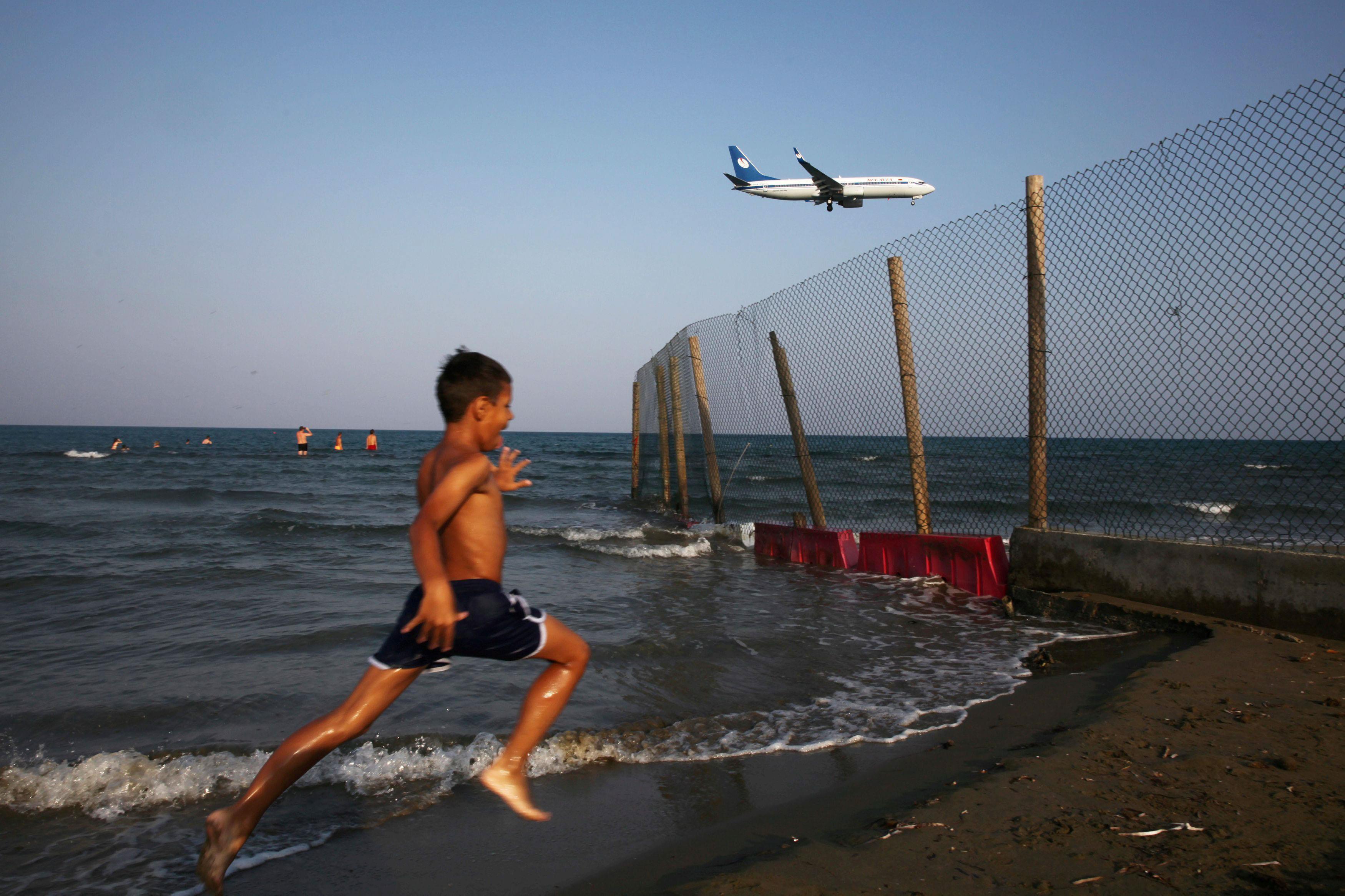 A boy runs on a beach while an airplane prepares to land at Larnaca International Airport