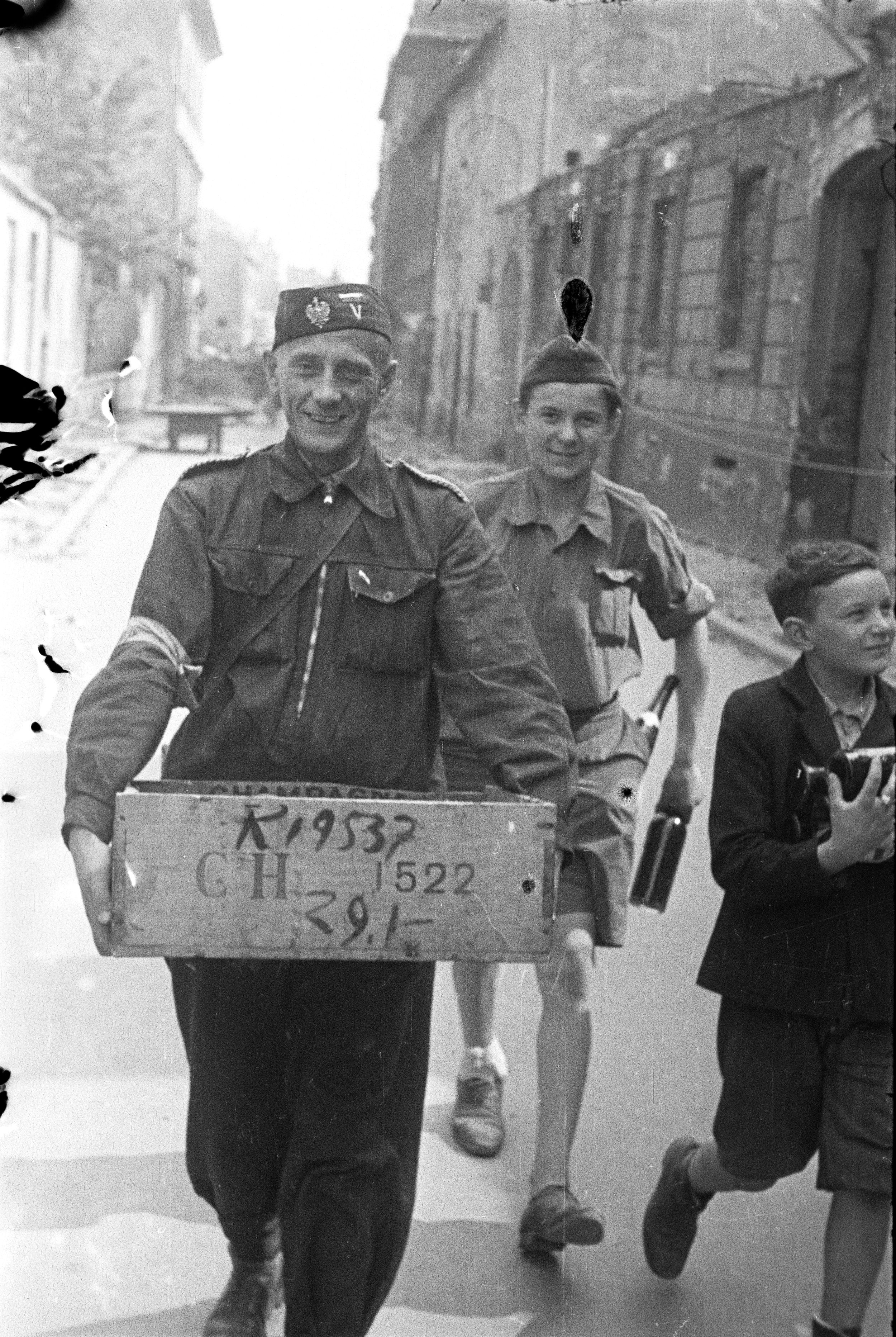 Ul. Warecka, widok w kierunku Nowego Światu. Na pierwszym planie grupa powstańców idących środkiem ulicy. Z lewej starszy mężczyzna w bluzie mundurowej spiętej pasem z koalicyjką, z biało-czerwoną opaską na prawym rękawie; na głowie ma furażerkę z orłem i biało-czerwoną naszywką. W rękach trzyma podłużną skrzynkę drewnianą z napisem