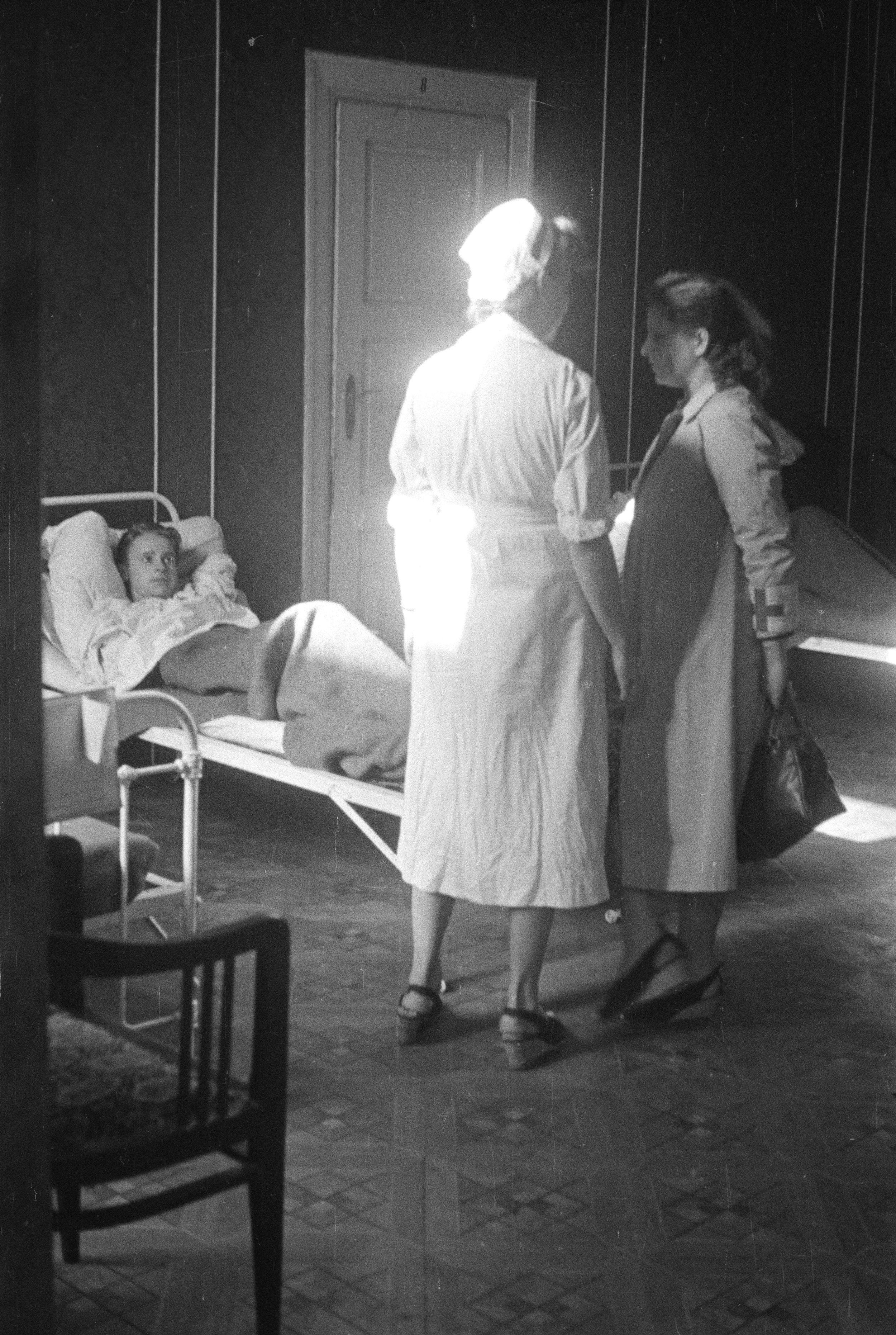 Szpital polowy przy ul. Konopczyńskiego 3. Na pierwszym planie dwie rozmawiające ze sobą sanitariuszki w fartuchach; kobieta po lewej ma na głowie czepek pielęgniarski, druga, z dużą torebką w ręku, ma na lewym rękawie opaskę z krzyżem. Na drugim planie okryty kocem powstaniec, leżący na żelaznym szpitalnym łóżku. Po lewej drugie łóżko (widoczny tylko mały fragment). W głębi zamknięte drzwi, w lewym dolnym rogu kadru wyściełane krzesło. Data: 2 - 6 sierpnia 1944r.