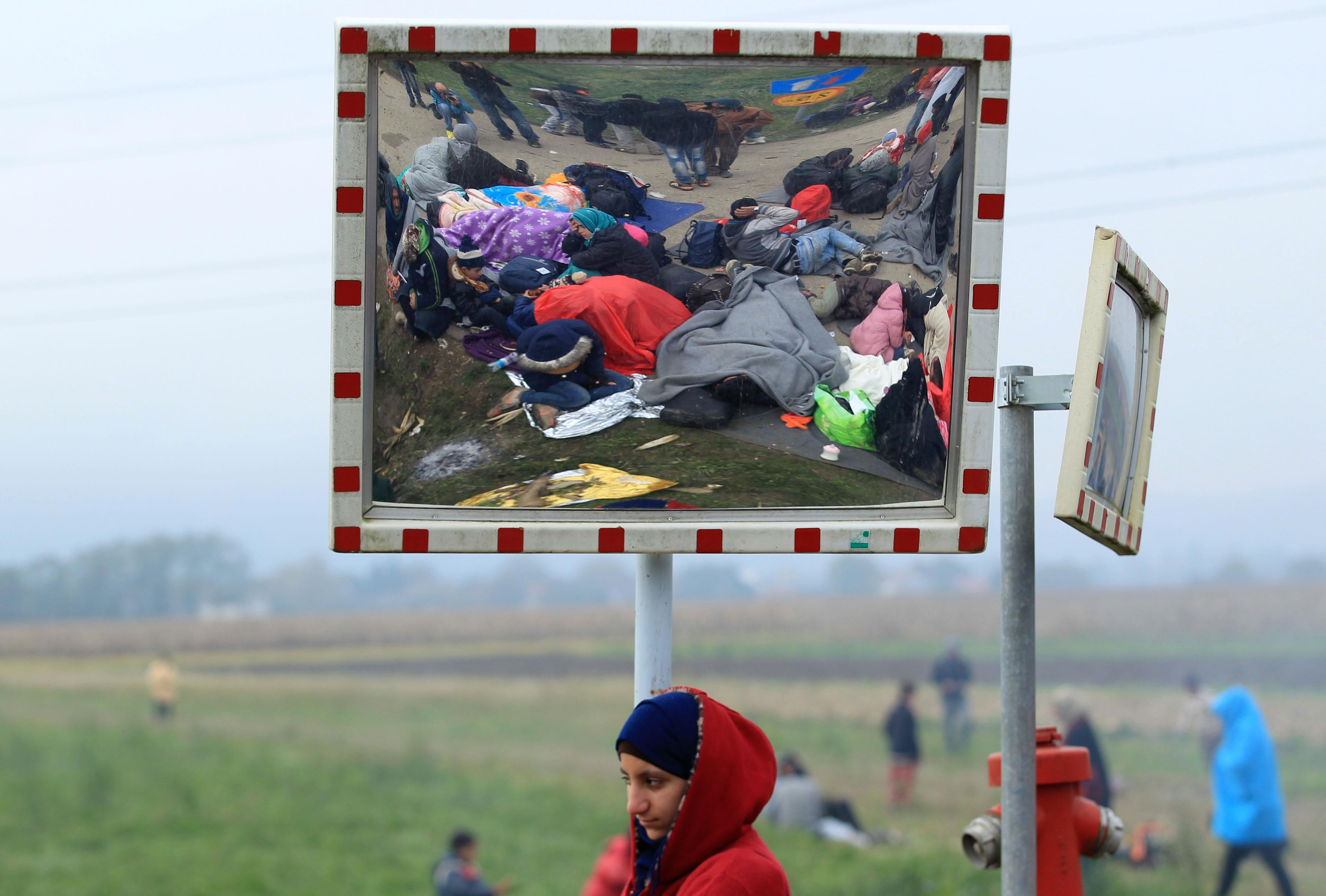 A girl looks on as resting migrants are seen in a street mirror after crossing the border from Croat