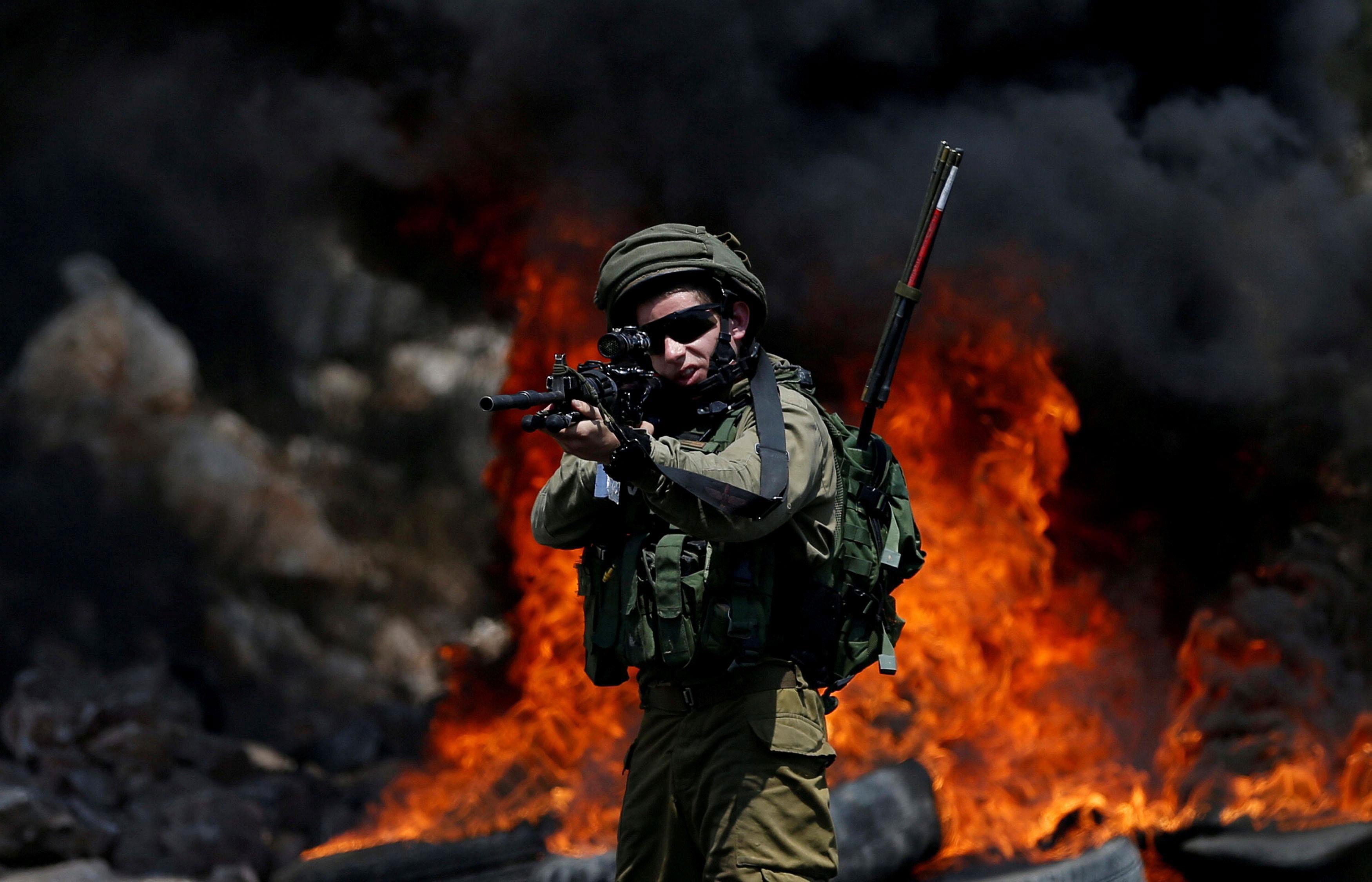 An Israeli soldier walks points his weapon toward Palestinian protesters during clashes in the West 