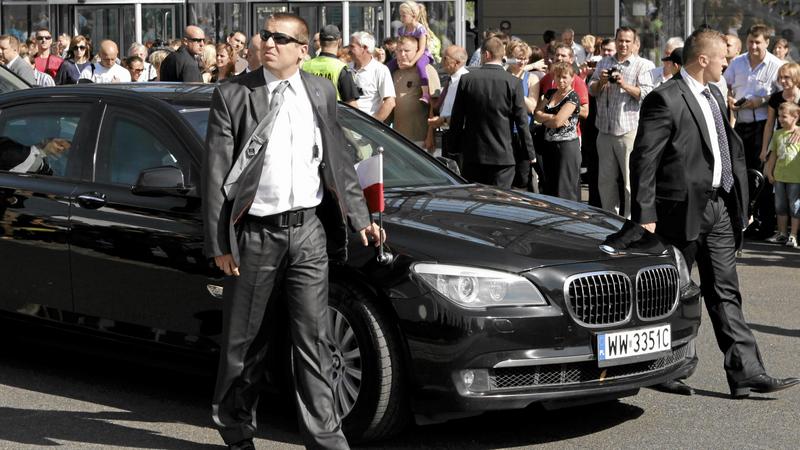 Ochroniarze w ciemnych okularach idą obok czarnego samochodu z polską flagą