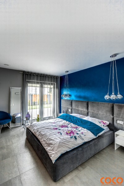 Sufitowe oświetlenie przestrzeni łóżka, wielki tapicerowany zagłówek oraz wykończona brokatem zagłówkowa szafirowa ściana nadają wnętrzu wrażenie luksusu.