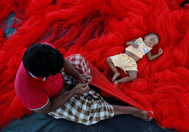 A fisherman stitches nets as his child sleeps beside him along the sea coast on the outskirts of Koc