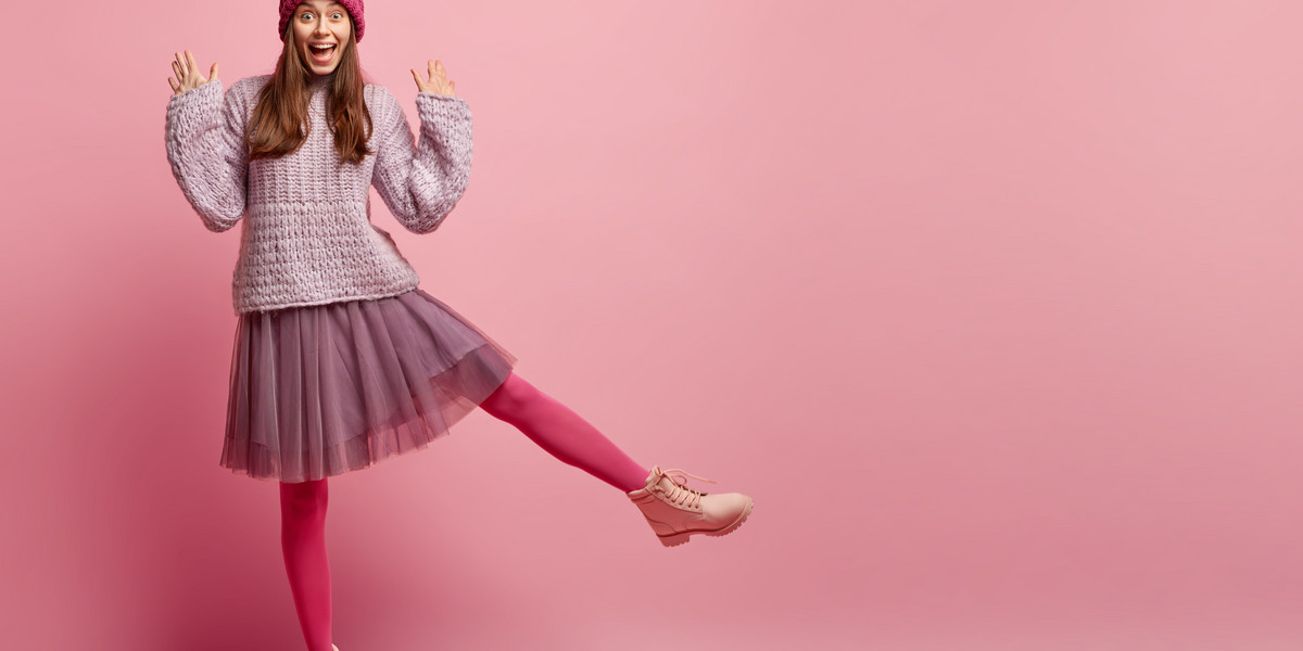 Studio shot of happy woman being in high spirit, shows palm, wears stylish clothes, raises leg, pose