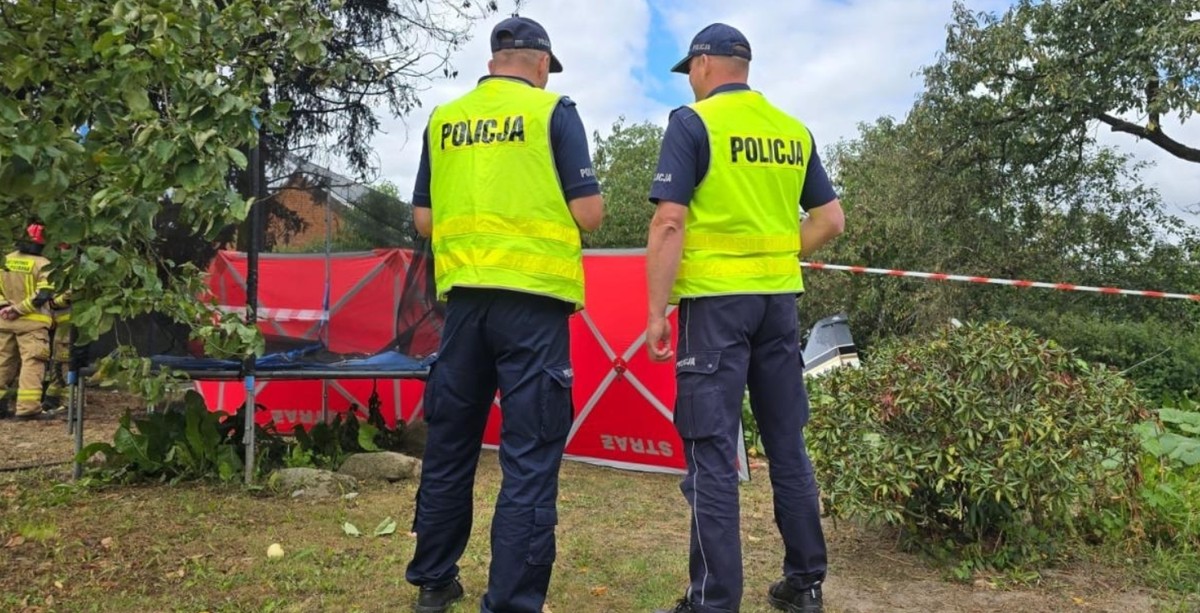 Dwóch policjantów w kamizelkach odblaskowych stoi tyłem, zabezpieczając miejsce wypadku. Obok widoczny jest strażak oraz czerwony parawan zasłaniający teren zdarzenia.