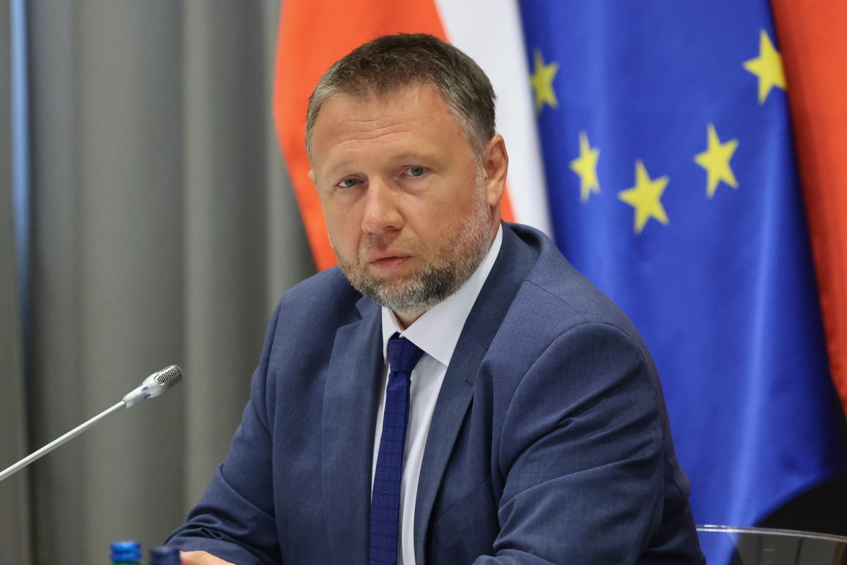 Zdjęcie Marcina Kierwińskiego, ministra spraw wewnętrznych i administracji, ubranego w granatowy garnitur, białą koszulę i krawat. Polityk siedzi przy stole z mikrofonem, na tle flagi Polski i Unii Europejskiej.
