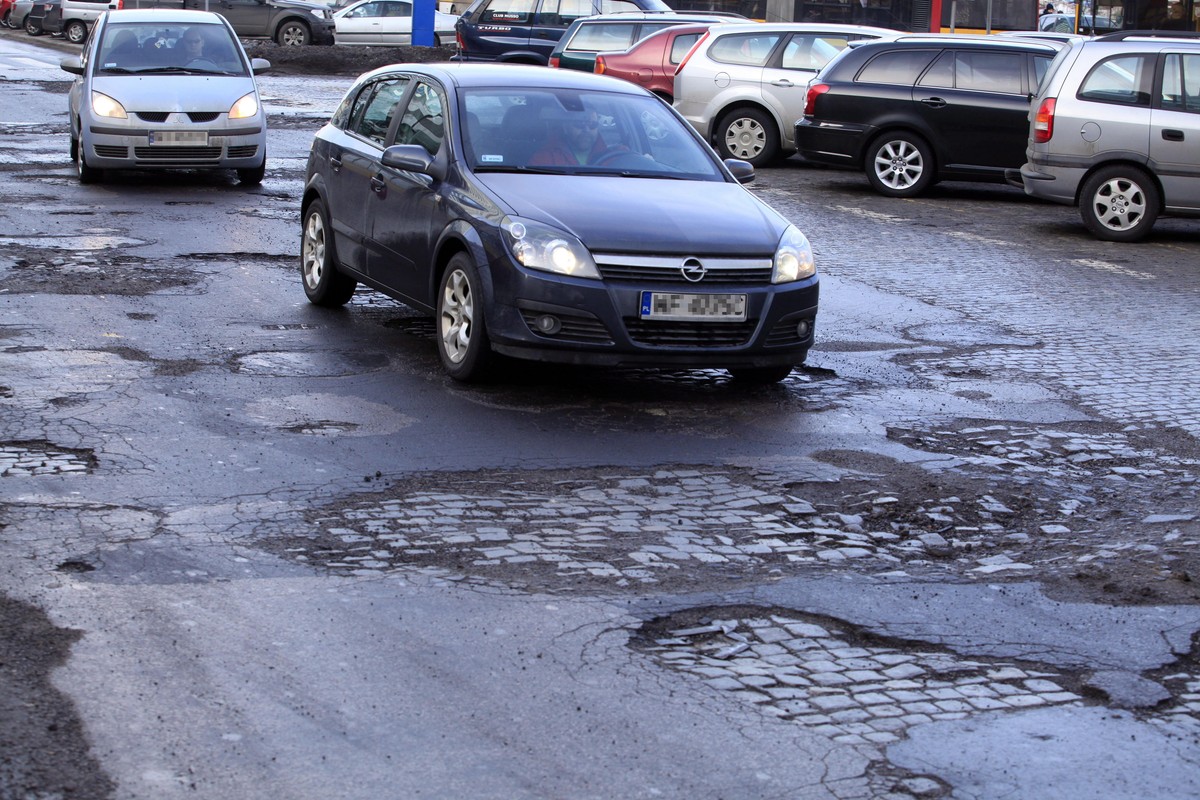 Samochody jadą po zniszczonej, pełnej dziur i pęknięć nawierzchni ulicy w centrum miasta; na poboczu widać zaparkowane pojazdy.
