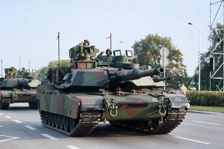 Na zdjęciu widać czołg Abrams w kamuflażu, podczas próby generalnej przed defiladą z okazji Święta Wojska Polskiego 15 sierpnia. Na wieży stoją żołnierze w hełmach, salutujący w stronę publiczności, a w tle widać inne pojazdy pancerne.