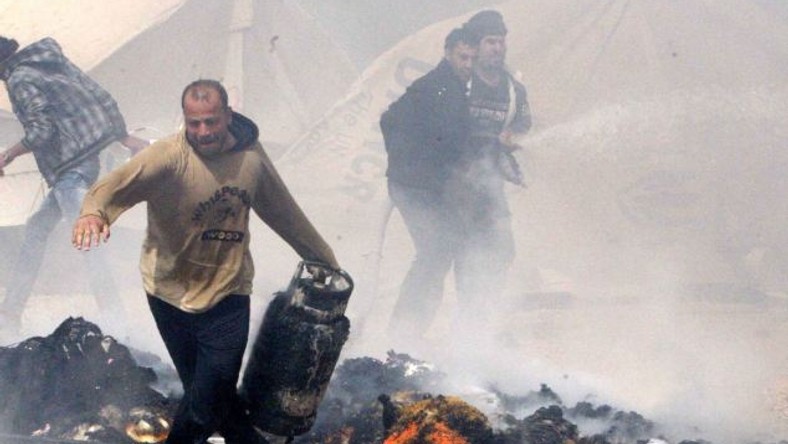 A Syrian refugee runs with a gas bottle cylinder, between burning tents.