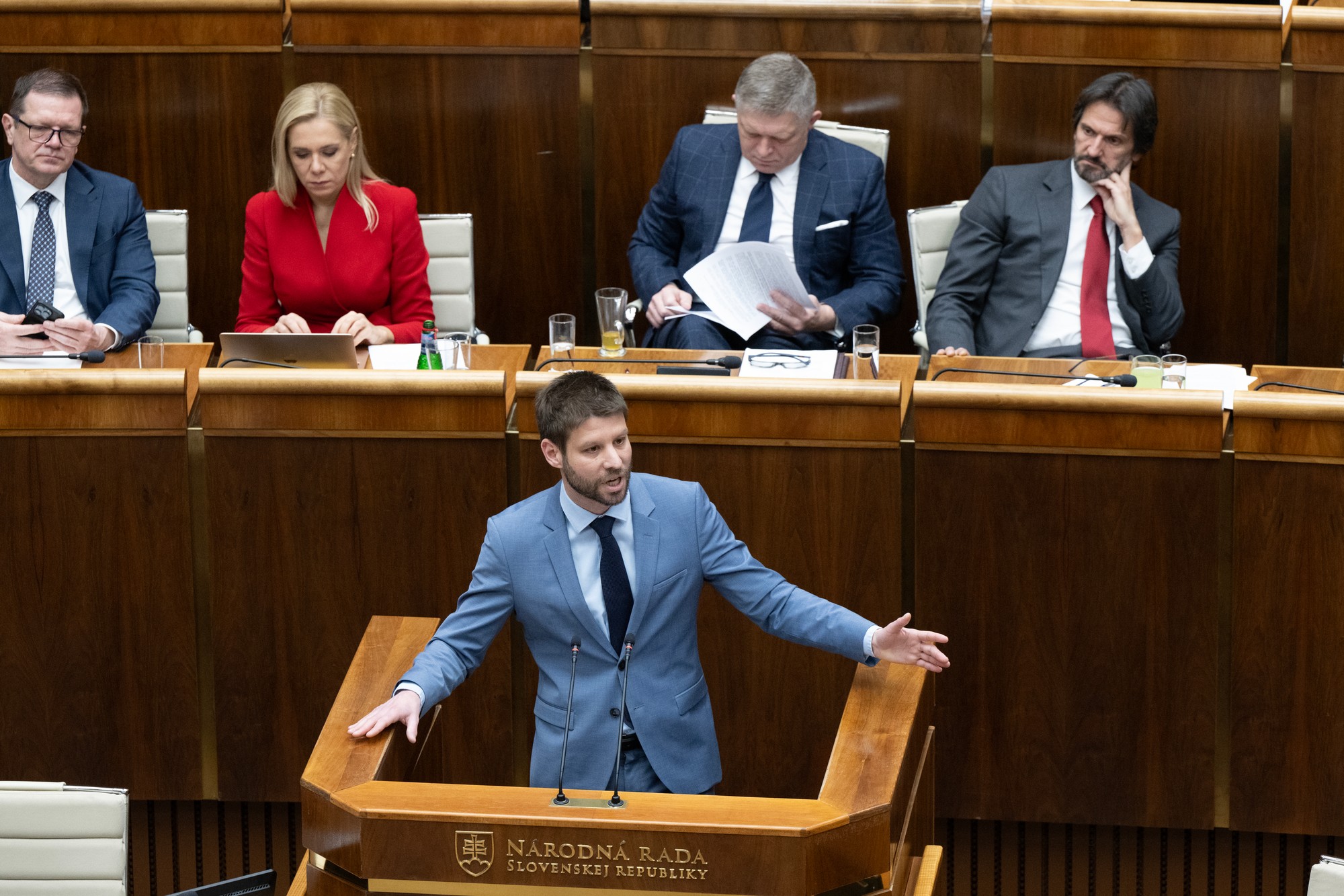 Uprostred poslanec NR SR Michal Šimečka (Progresívne Slovensko) hovorí počas mimoriadnej schôdze. V pozadí zľava podpredseda vlády SR pre Plán obnovy a odolnosti a využívanie eurofondov Peter Kmec (Hlas-SD), podpredsedníčka vlády a ministerka hospodárstva SR Denisa Saková (Hlas-SD), premiér Robert Fico, vicepremiér a minister obrany SR Robert Kaliňák (obaja Smer-SD).