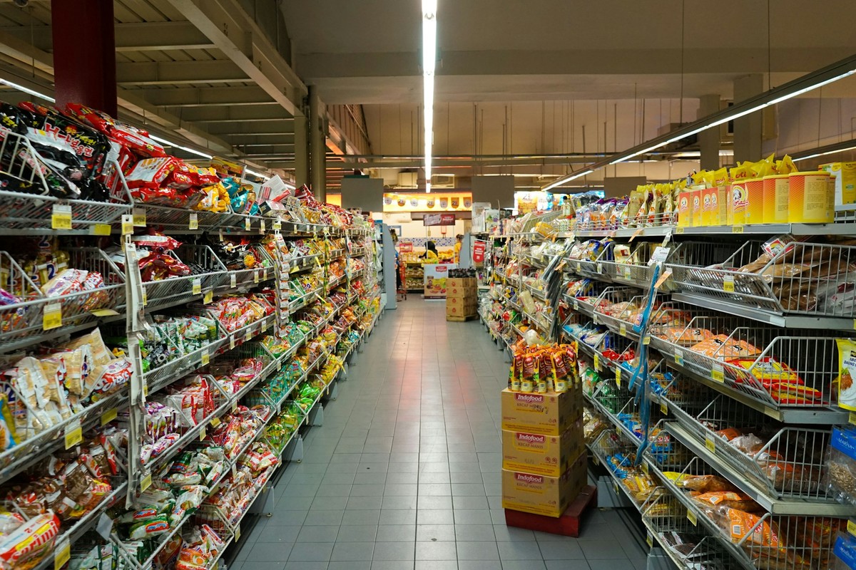 Alejka w supermarkecie z półkami wypełnionymi różnorodnymi produktami spożywczymi w kolorowych opakowaniach; pośrodku stoi stos kartonów z makaronem błyskawicznym.