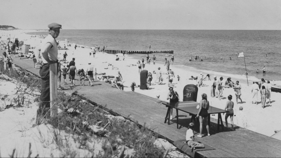 Plażowicze podczas wypoczynku. Widoczny drewniany deptak, wiklinowe kosze plażowe oraz stół do ping-ponga.