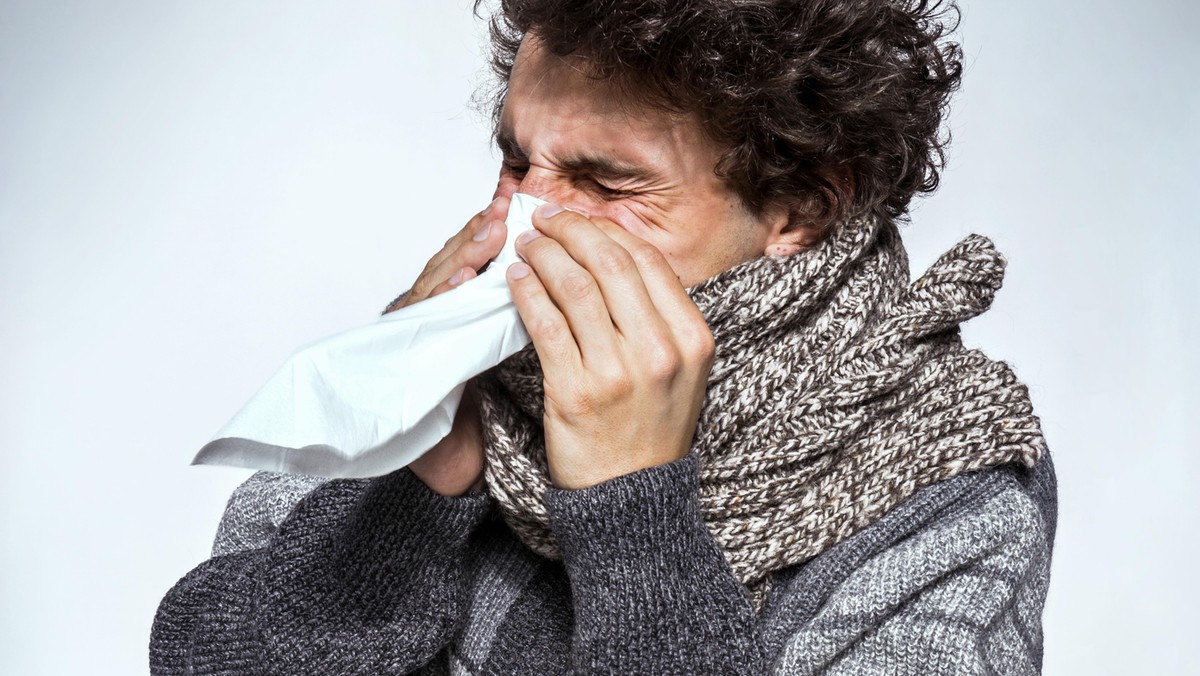 Ill young man with red nose, scarf and cap sneezing into handkerchief