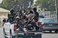 Soldiers and people carrying machetes ride on the back of a vehicle along a street in Gombe