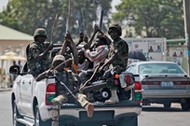 Soldiers and people carrying machetes ride on the back of a vehicle along a street in Gombe