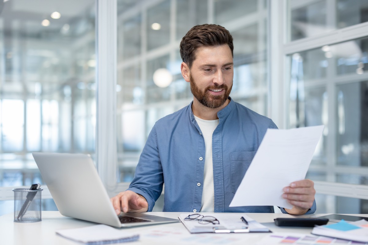 Young,Businessman,Smiling,,Holding,Paperwork,While,Typing,On,Laptop,Computer,