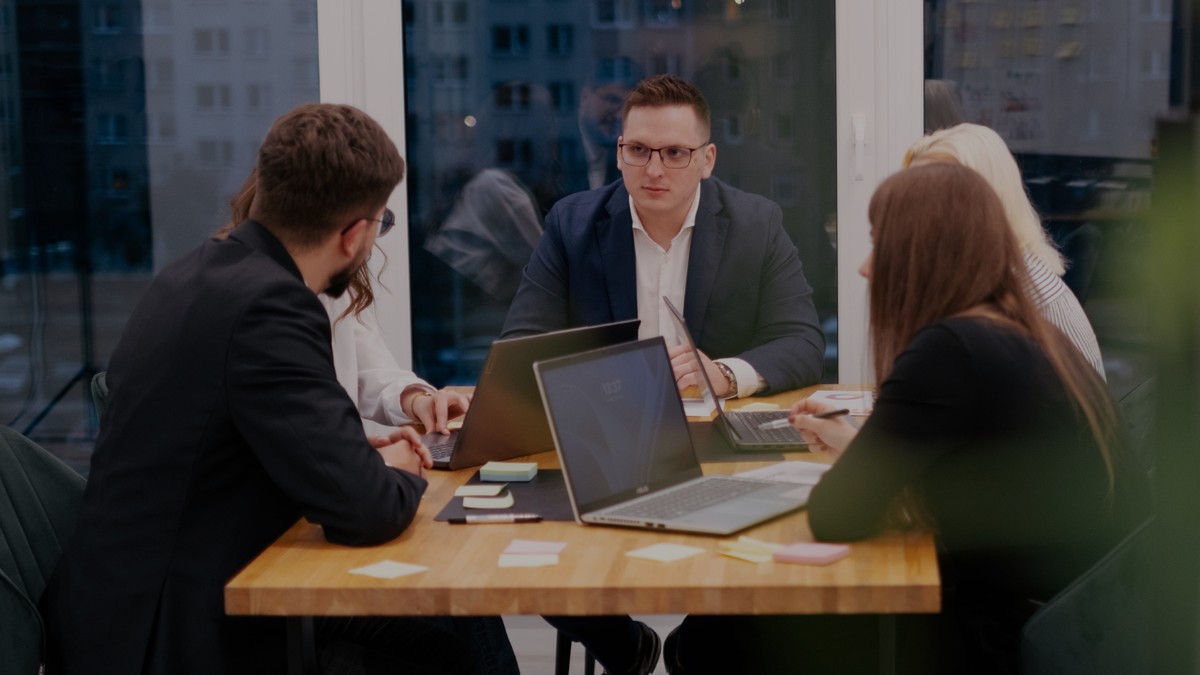 Grupa osób w biurze siedzi przy stole z laptopami i dokumentami, prowadząc rozmowę podczas spotkania biznesowego.
