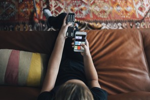 High angle view of teenage girl using phone app and remote control while sitting on sofa watching TV