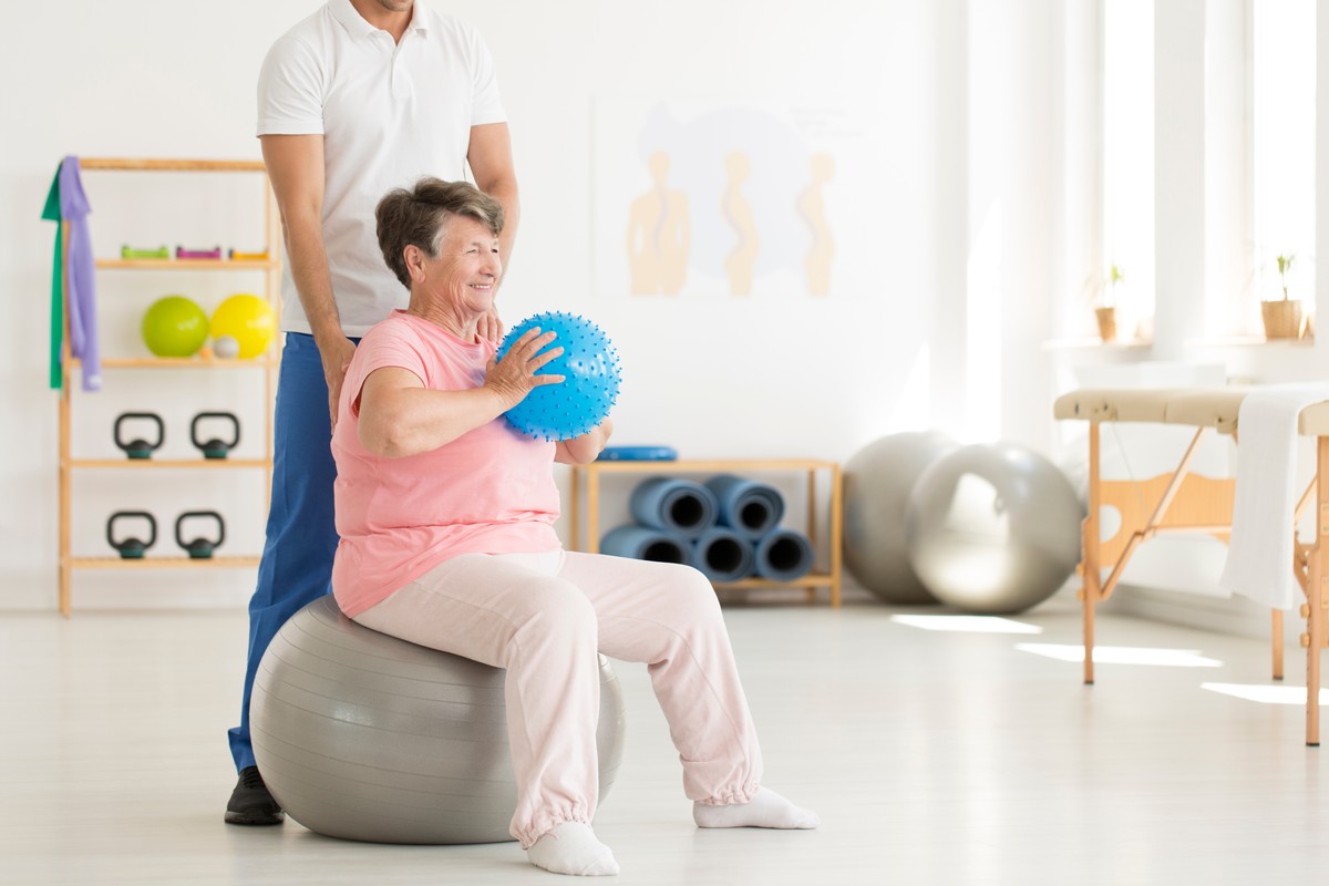 Sanatorium senior seniorka lekarz uzdrowisko  trening ćwiczenia rehabilitacja Happy,Senior,Woman,Sitting,On,Grey,Ball,And,Holding,Blue
