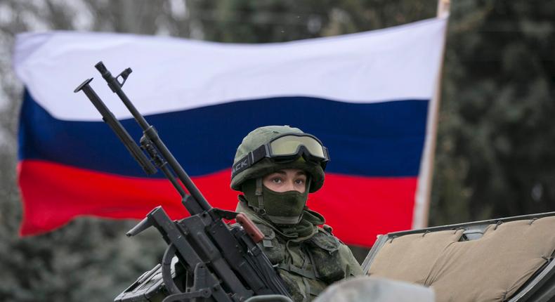 A pro-Russian man (not seen) holds a Russian flag behind an armed serviceman on top of a Russian army vehicle outside a Ukrainian border guard post.