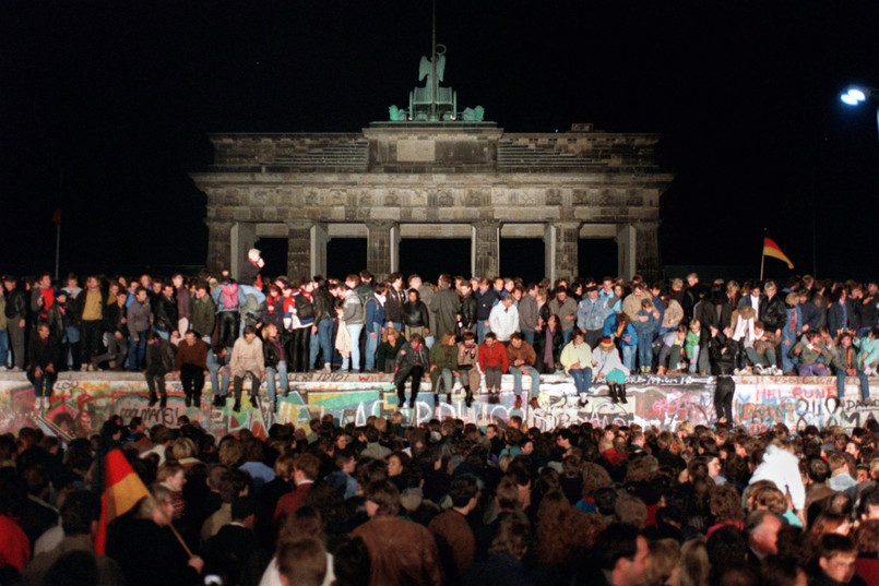 Zdjęcie z 10 listopada 1989. Berlińczycy świętują otwarcie granic przed Bramą Brandenburską w Berlinie. Mur berliński był systemem umocnień o długości ok. 156 km (betonowy mur, okopy, zapory drutowe, miny). W języku enerdowskiej propagandy nazwany antyfaszystowskim wałem ochronnym. Funkcjonował od 13 sierpnia 1961 do 9 listopada 1989 oddzielając Berlin Zachodni od Wschodniego. Padł w nocy z czwartku 9 listopada na piątek 10 listopada 1989, po przeszło 28 latach istnienia. Był on jednym z najbardziej znanych symboli zimnej wojny i podziału Niemiec. Podczas prób przedostania się przez strzeżone urządzenia graniczne do Berlina Zachodniego wielu ludzi zostało zabitych. Różne źródła podają od 136 do 238 śmiertelnych przypadków.