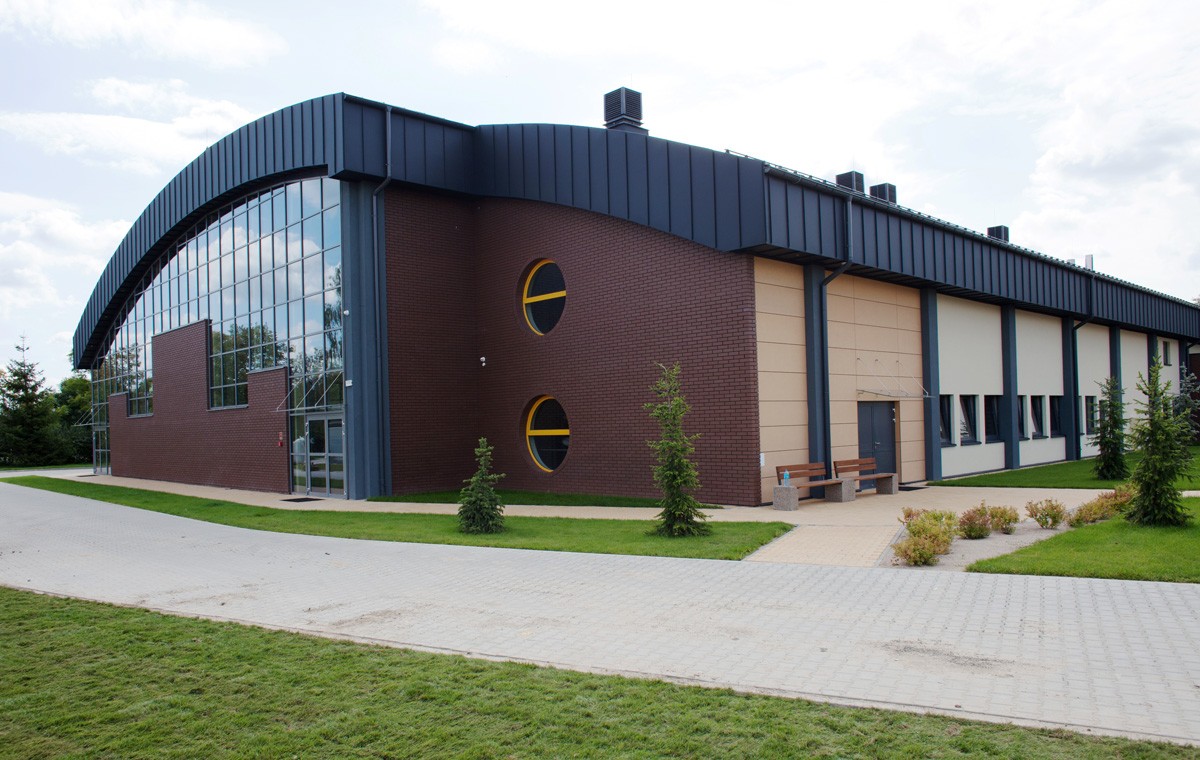 Nowoczesny budynek hali sportowej z dużymi przeszkleniami i charakterystycznym łukowatym dachem, otoczony chodnikiem i zielenią.