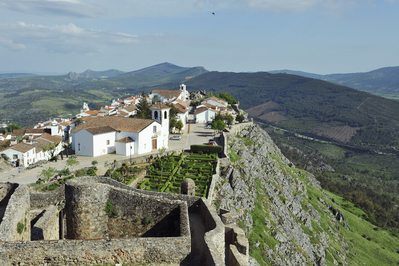 Marvão Zaledwie dwie i pół godziny jazdy od Lizbony, w wąwozie niedaleko granicy z Hiszpanią, znajduje się niewielka osada otoczona murami. To najwyżej położona wioska w Portugalii, którą zwykło określać się jako gniazdo orła ukryte w górach. Marvão, z panoramicznym widokiem na rozległe równiny regionu Alentejo, to jedno z najładniejszych miejsc w całej południowej Europie. Koniecznie trzeba zobaczyć tutejszy zachód słońca – światło odbijające się od kwarcu w granitowych głazach, które podtrzymują ogromne mury zamku, daje żywe, niemal jarzące się kolory, a także słynne fortyfikacje. – Pierwsze fortyfikacje wzniesione zostały tutaj przez Maurów w ósmym wieku. Do dziś XIII-wieczne mury zachowały się praktycznie nietknięte. Do osady prowadzi z nich wąskie, średniowieczne przejście, przy którym znajduje się ciekawie ukształtowany mauretański budynek zwany kaplicą jerozolimską. Jego wnętrze przez setki lat nie było modernizowane, nie zmieniło się tam nic. Na szczycie znajduje się piękna, niska dzwonnica z maleńką kopułą. Wyjątkowym elementem jest także piękna drewniana galeria, a na jednym z bocznych ołtarzy znajduje się niezwykły posąg Trójcy Przenajświętszej – mówi Piotr Wilk z biura podróży Rainbow. Z twierdzy zamkowej widać całą okolicę – leżące na południu Serra de São Mamede i urocze miasto Estremoz, na północy góry Serra da Estrela (góry Gwiazd) z Castelo de Vide na północnym zachodzie, a na północnym wschodzie, za horyzontem roztacza się już Hiszpania. Źródło: r.pl/portugalia >>>