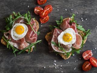 Sandwich with prosciutto, arugula, and fried quail eggs on wooden background, top view. Tasty breakfast, snack or appetizer