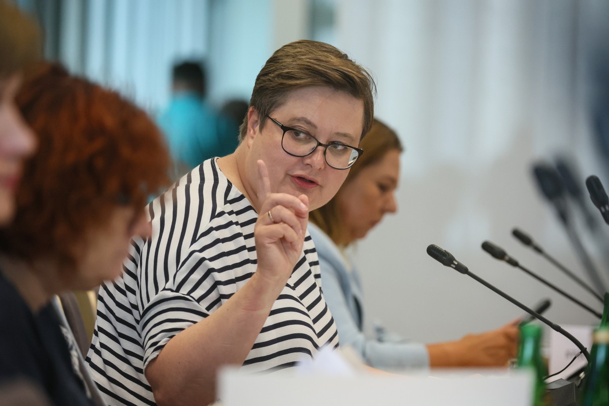 Wiceminister edukacji narodowej Katarzyna Lubnauer ubrana w biało-czarną bluzkę w paski, siedzi przy stole konferencyjnym i mówi, unosząc palec wskazujący. Na stole stoją mikrofony i butelki z wodą, w tle widać inne uczestniczki spotkania.
