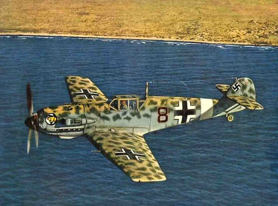 Niemiecki samolot, konkretnie Messerschmitt Bf 109E-4/Trop w trakcie przelotu nad północnoafrykańskim wybrzeżem (domena publiczna).