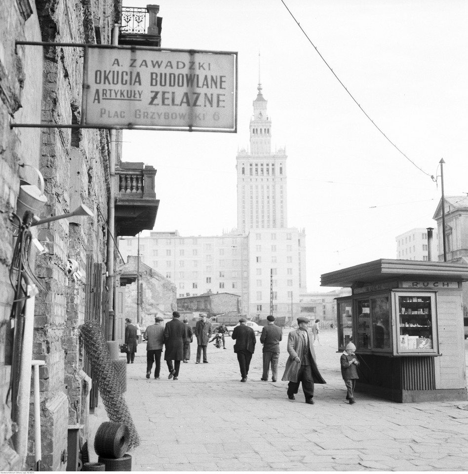 Plac Grzybowski w Warszawie. Fragment zabudowy placu od strony ul. Królewskiej. Z lewej widać zawieszoną tablicę z napisem "A. Zawadzki, okucia budowlane, artykuły żelazne, plac Grzybowski 6". Po prawej stoi kiosk "RUCH-u". Na dalszym planie widać wieżę pałacu Kultury i Nauki (1961 r.)