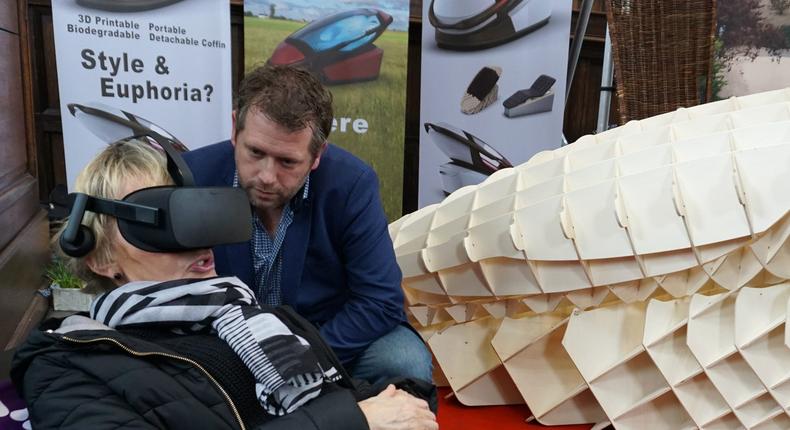 Dutch designer Alexander Bannink explains how the Sarco euthanasia pod works as a woman experiences sitting in the device by wearing virtual reality glasses, on April 14, 2018 at the Amsterdam Funeral Expo.