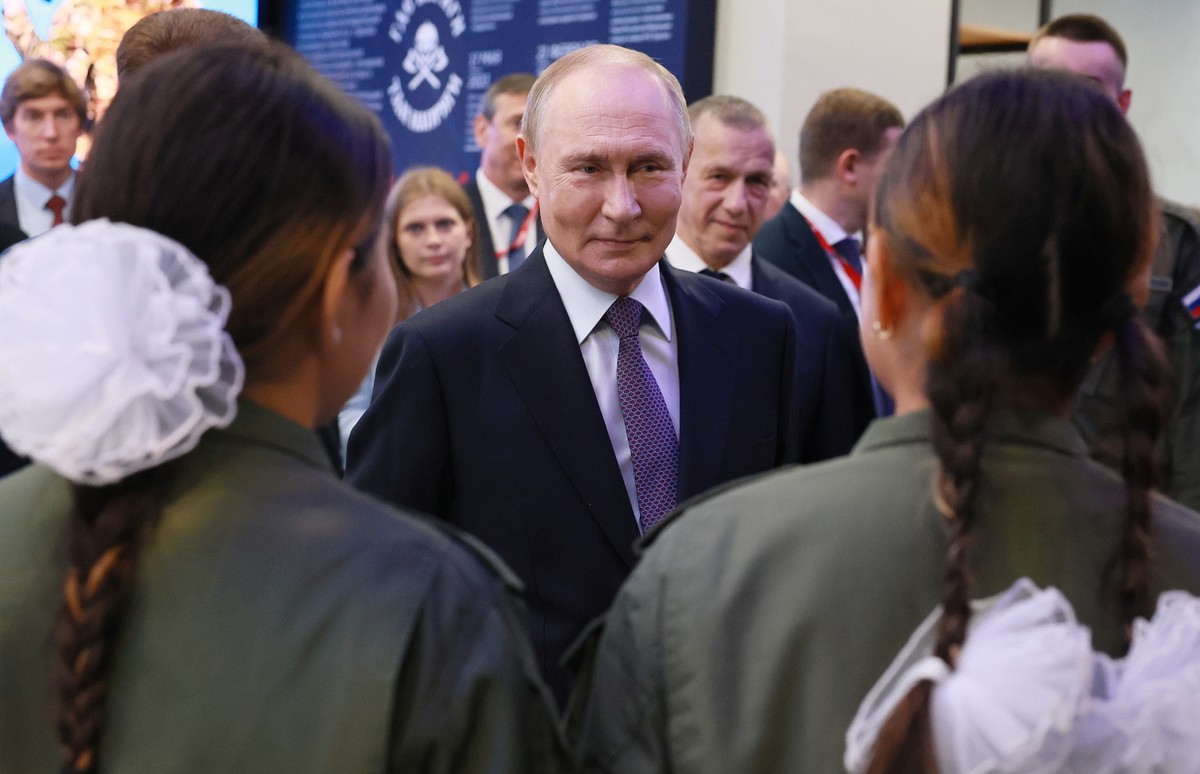 Prezydent Rosji Władimir Putin w garniturze i krawacie stoi w otoczeniu ludzi, patrząc w stronę dwóch dziewcząt w zielonych strojach z białymi kokardami we włosach. W tle widoczna jest grupa osób w formalnych ubraniach.