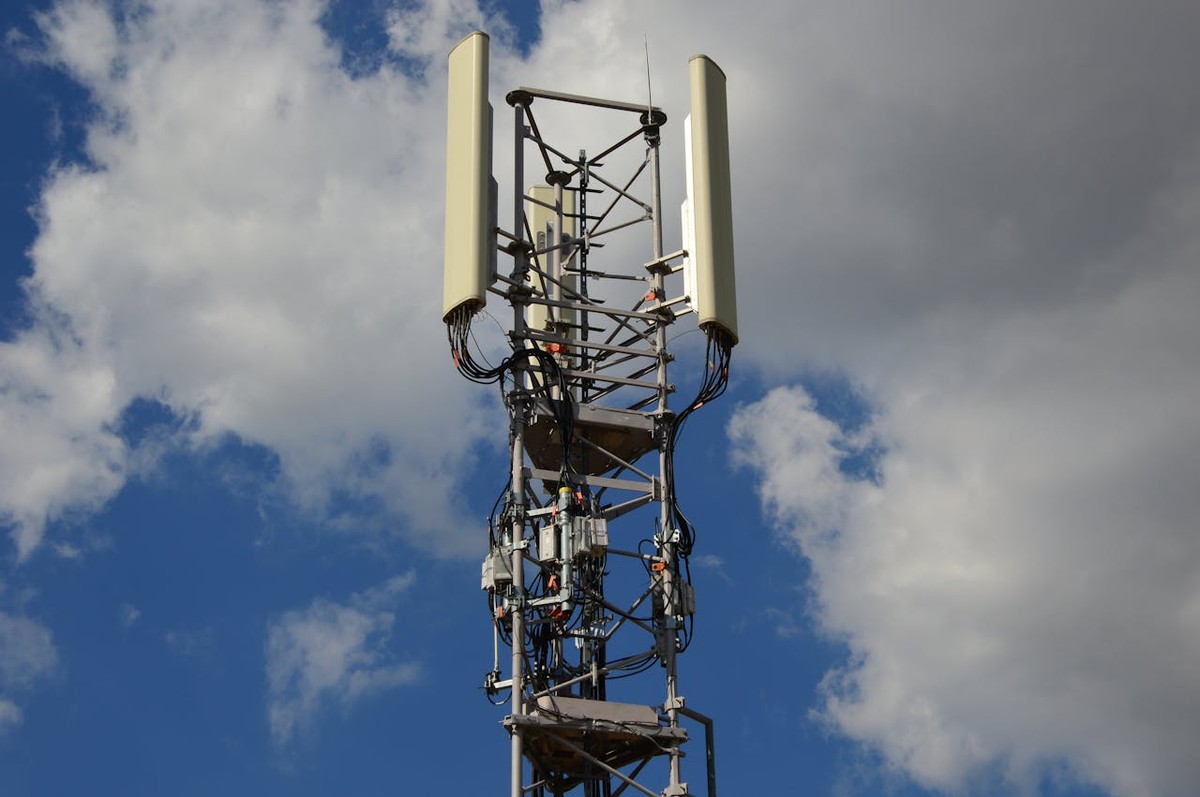 Maszt telekomunikacyjny z antenami i przewodami na tle nieba z białymi chmurami.