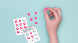 Top view of female hand holding pink prescription pill next to blister packs over pastel blue background. Sick patient taking medication.