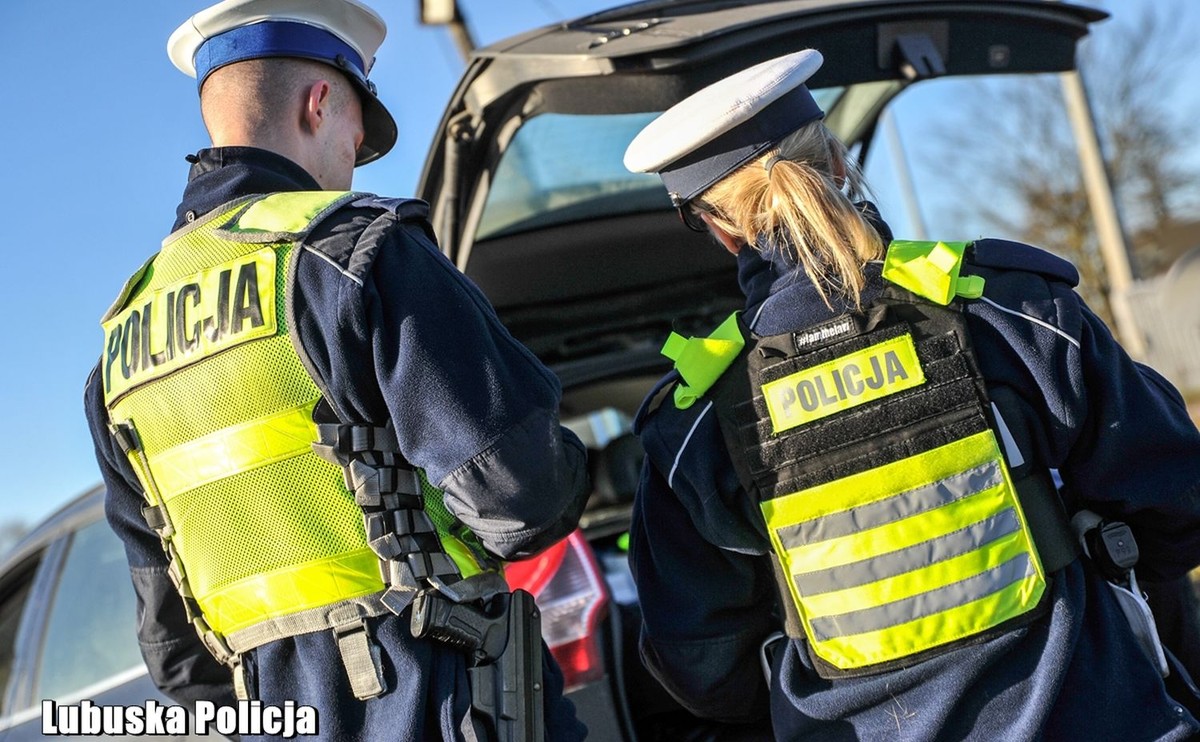 Dwoje policjantów w odblaskowych kamizelkach z napisem "POLICJA" sprawdza otwarty bagażnik zatrzymanego do kontroli samochodu osobowego