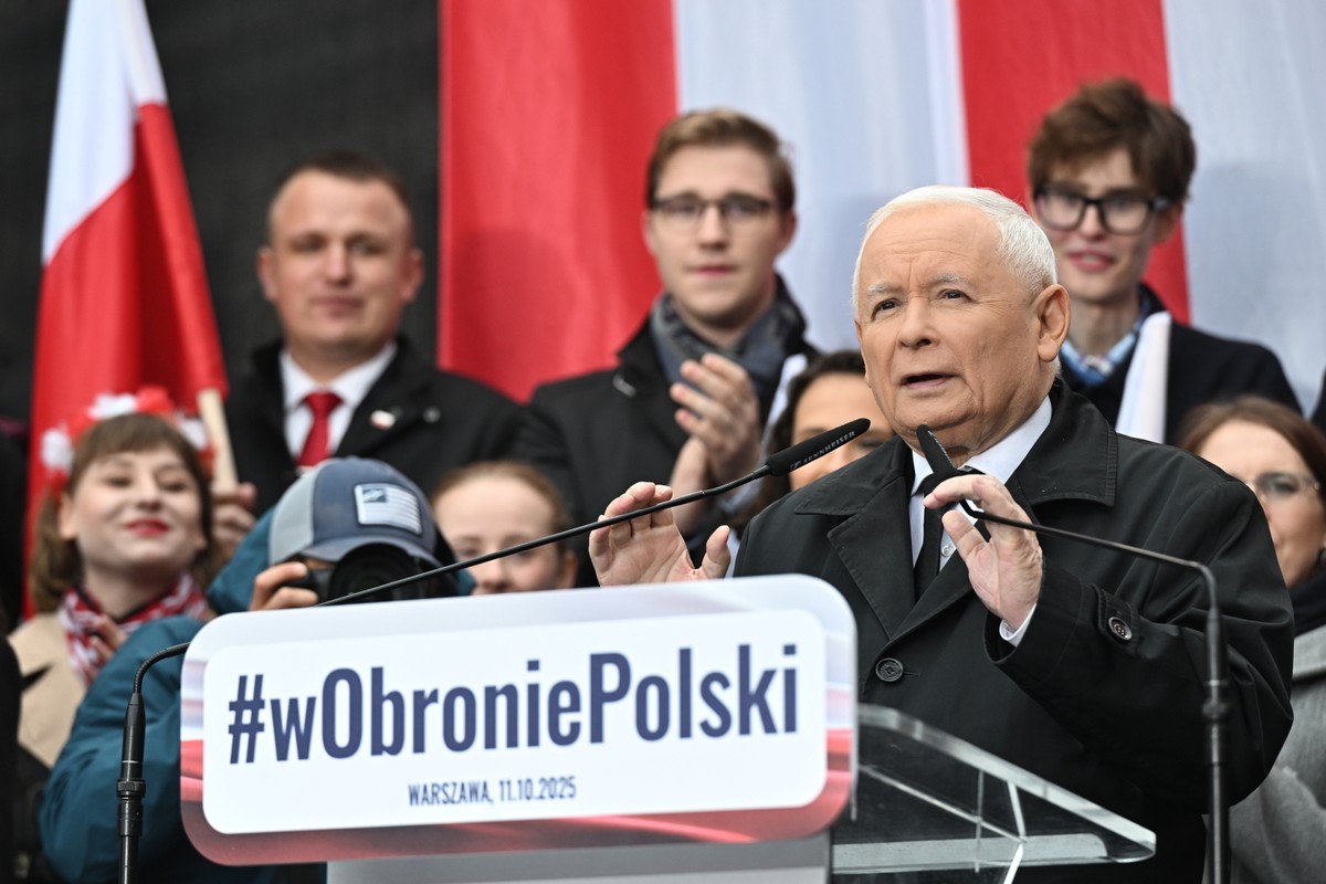 Jarosław Kaczyński przemawia na wiecu przy mównicy z napisem „#wObroniePolski”, w tle stoją ludzie z biało-czerwonymi flagami.