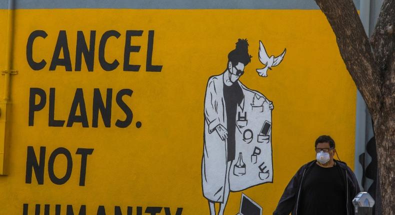 A man wearing gloves and a face mask walks by a mural reading Cancel Plans Not Humanity in Los Angeles, California