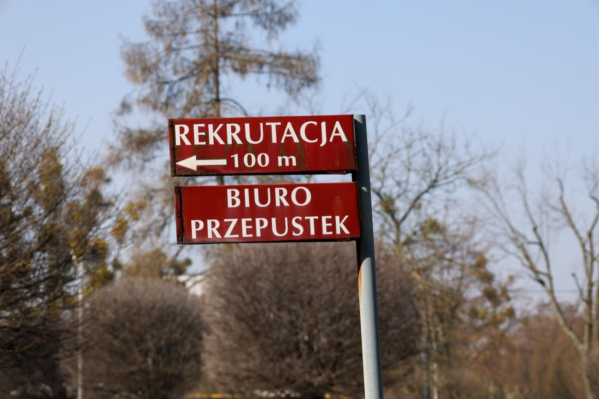 Czerwone tablice informacyjne przytwierdzone do metalowego słupa. Na górnej tablicy napis „REKRUTACJA 100 m” z białą strzałką w lewo, a na dolnej napis „BIURO PRZEPUSTEK”. W tle widać nagie drzewa i błękitne niebo.