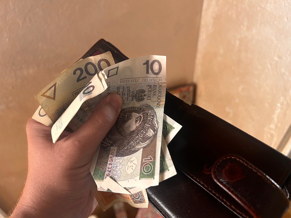 Dłoń trzymająca portfel oraz kilka banknotów polskich złotych, w tym widoczne nominały 200, 10 i 20 złotych.