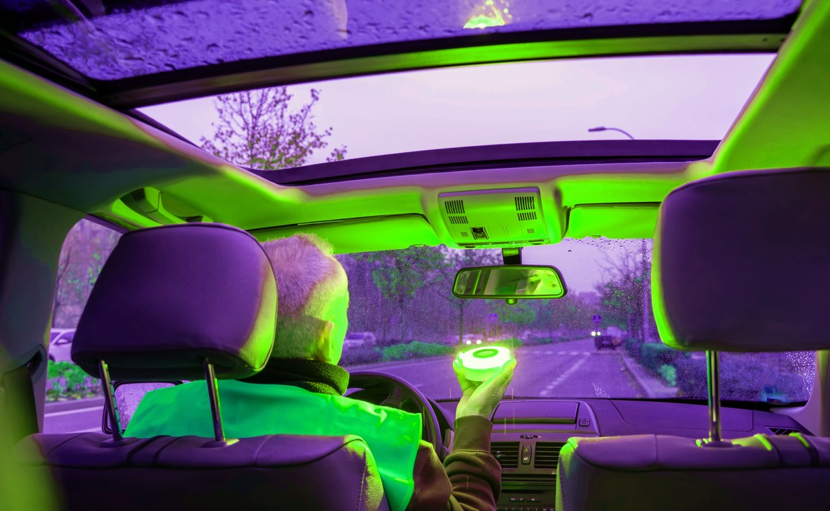 Widok z wnętrza samochodu na kierowcę w żółtej kamizelce odblaskowej, który trzyma w dłoni świecący na zielono, okrągły sygnalizator błyskowy na tle przedniej szyby i drogi po zmroku