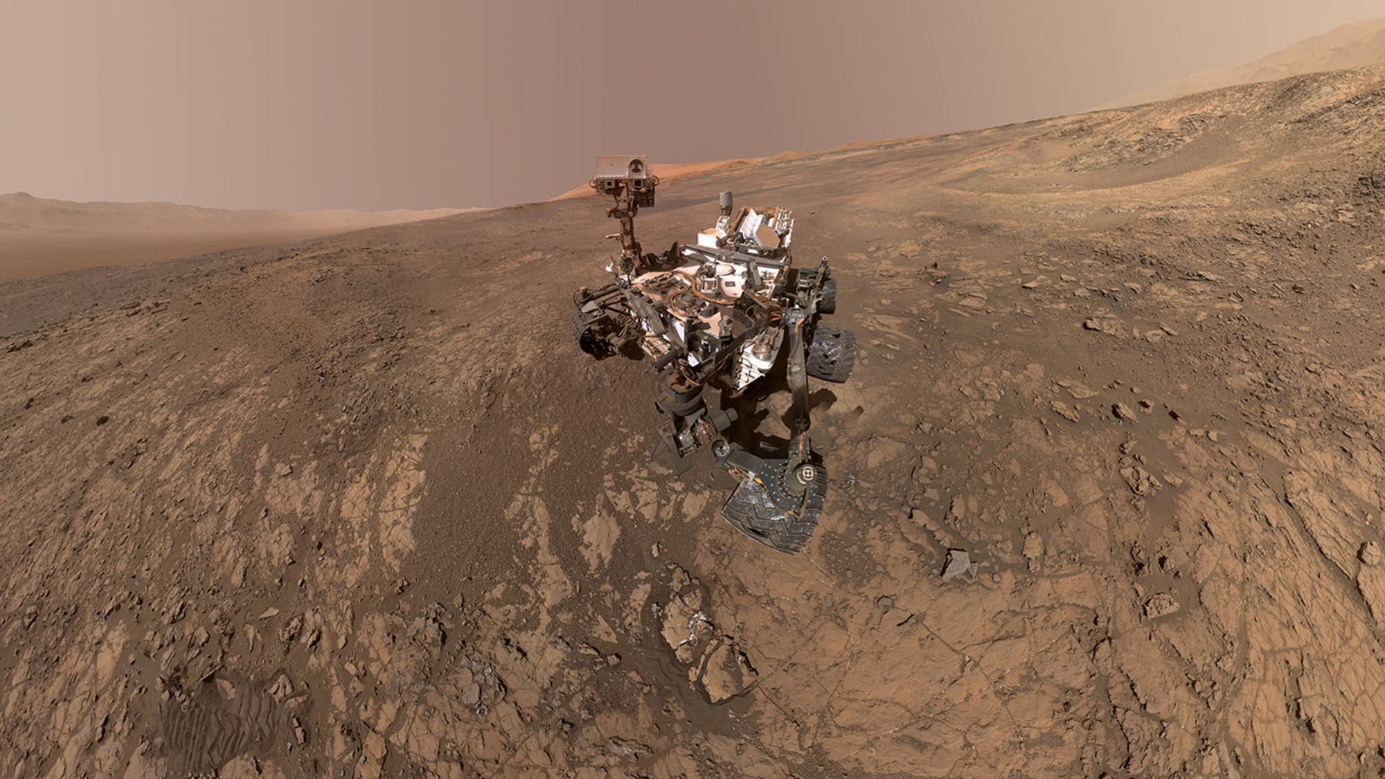 Sonda Curiosity s okrajom krátera Gale na horizonte. Ostrá hora vytŕčajúca spoza sondy je Mount Sharp.