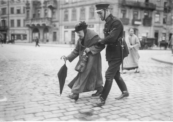 Policjant przeprowadzający staruszkę przez ulicę Krakowskie Przemieście (1925, domena publiczna).