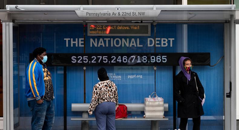 Passengers wearing face masks wait for their bus in front of a national debt display on Pennsylvania Ave. NW in Washington on Monday, May 18, 2020
