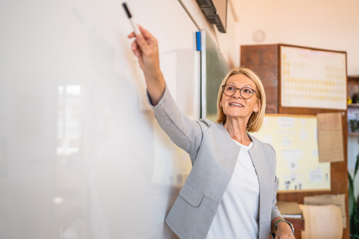 Mature,Woman,Teacher,Stand,At,Whiteboard,In,Classroom,,Teach,A