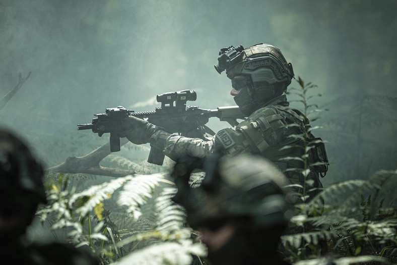 Fotografia przedstawiająca żołnierza ubranego w hełm HBT-02 oraz kamizelkę KBT-01. Dodatkowo na karabinku dostrzec możemy Celownik Kolimatorowy CK-3 i Powiększalnik Kolimatorowy PK-1.